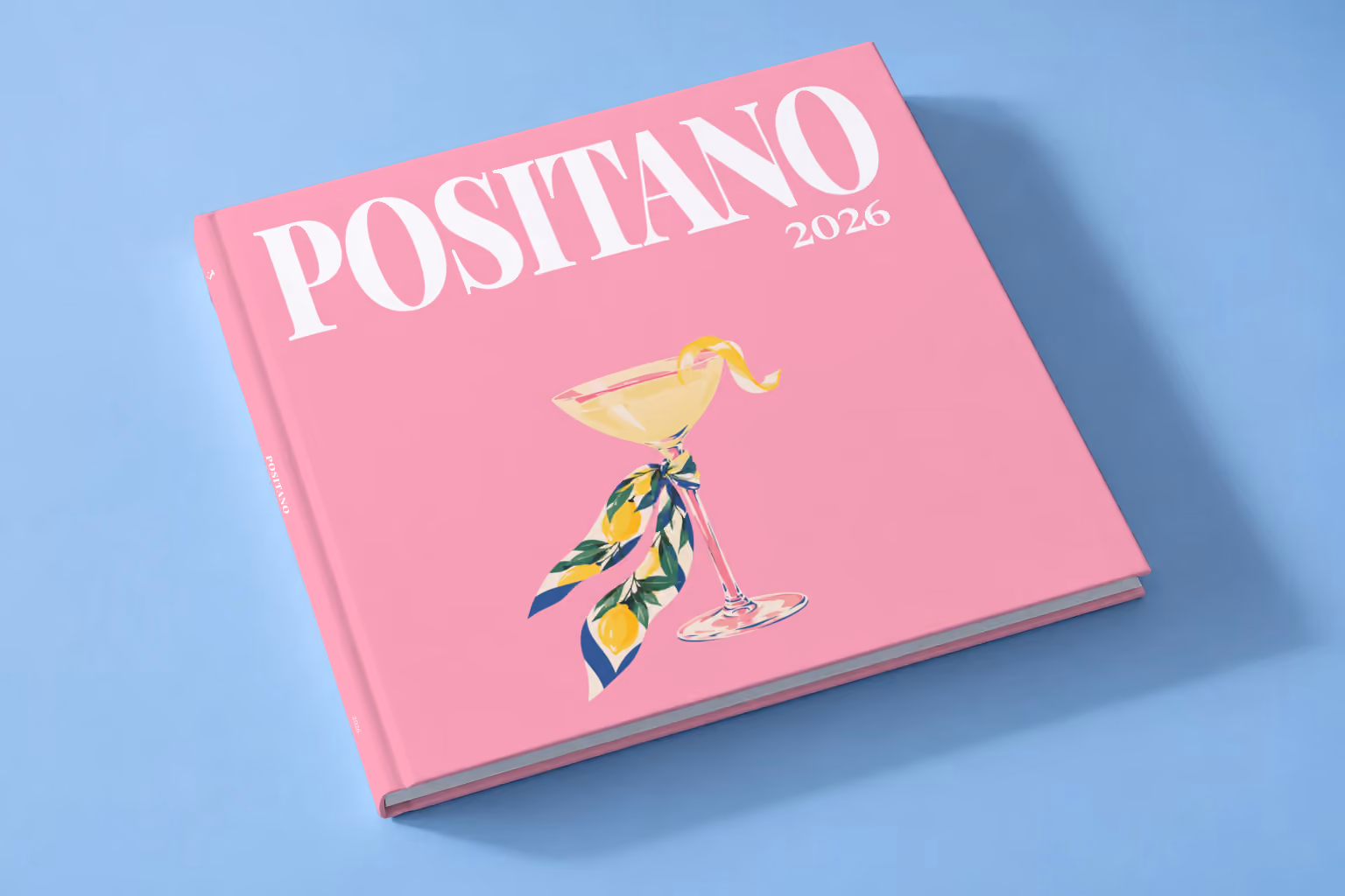 Italy square hardcover photo book with the Positano illustrated cover of a martini glass tied with a lemon-print scarf on pink, lying on a pale blue studio backdrop