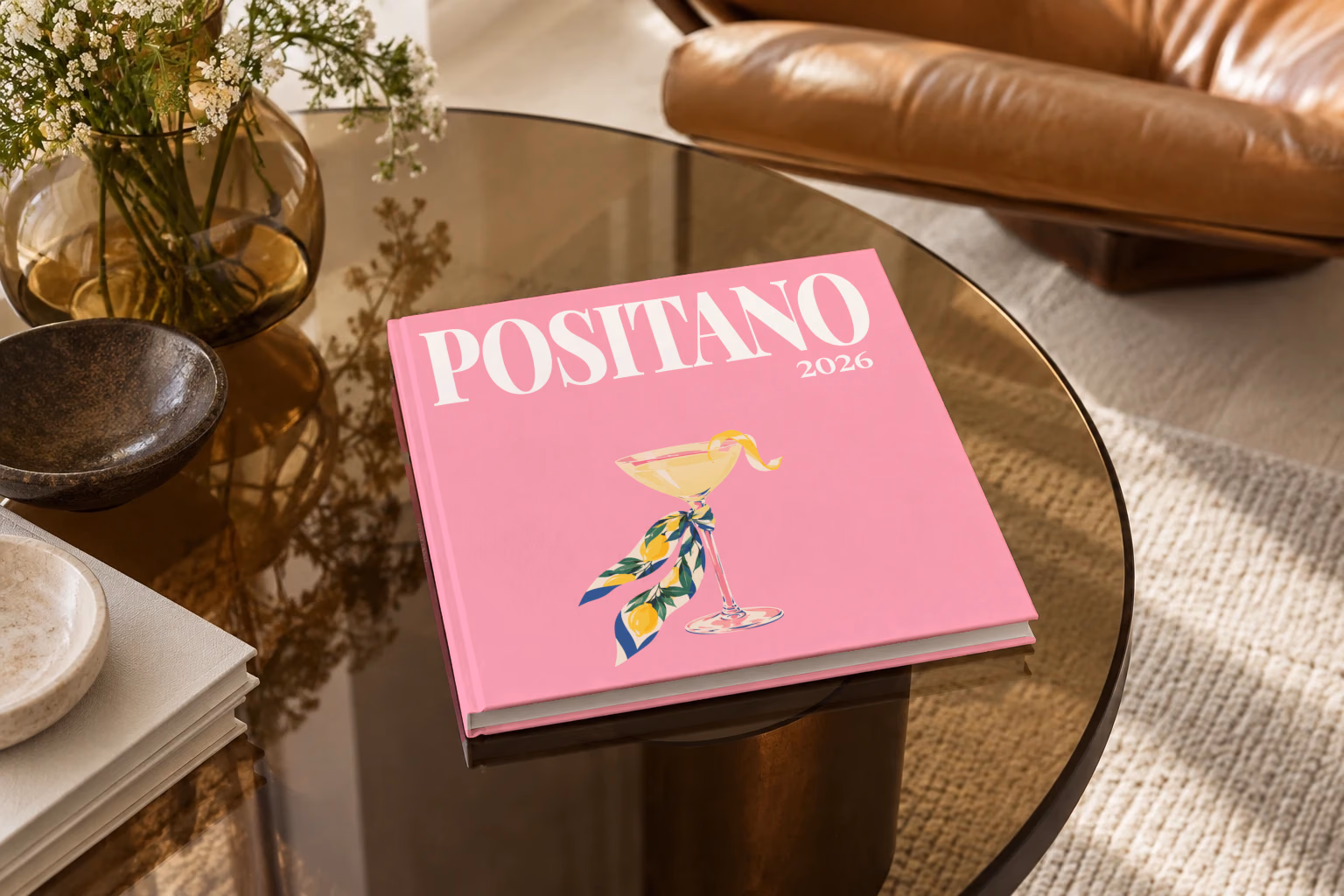 Italy square hardcover photo book with the Positano illustrated cover of a martini glass tied with a lemon-print scarf on pink, on a smoked glass coffee table beside a leather armchair in warm afternoon light