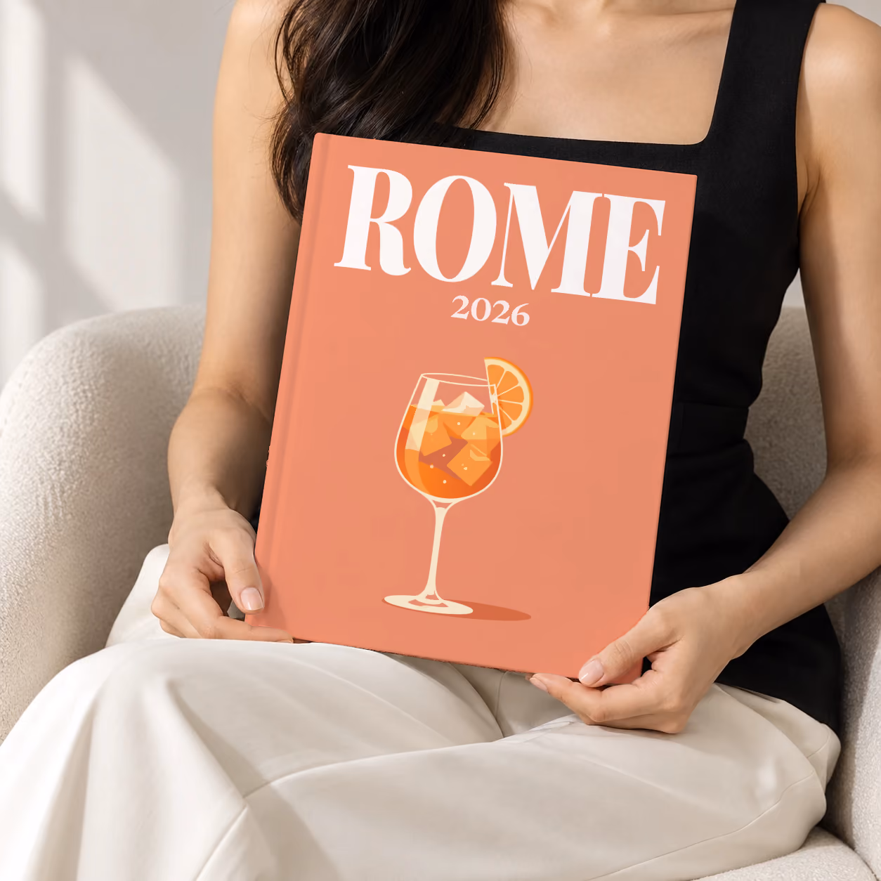 Italy hardcover photo book with the Rome illustrated cover of an Aperol spritz on coral, held upright in two hands by a woman in a black top, sitting in a cream boucle armchair