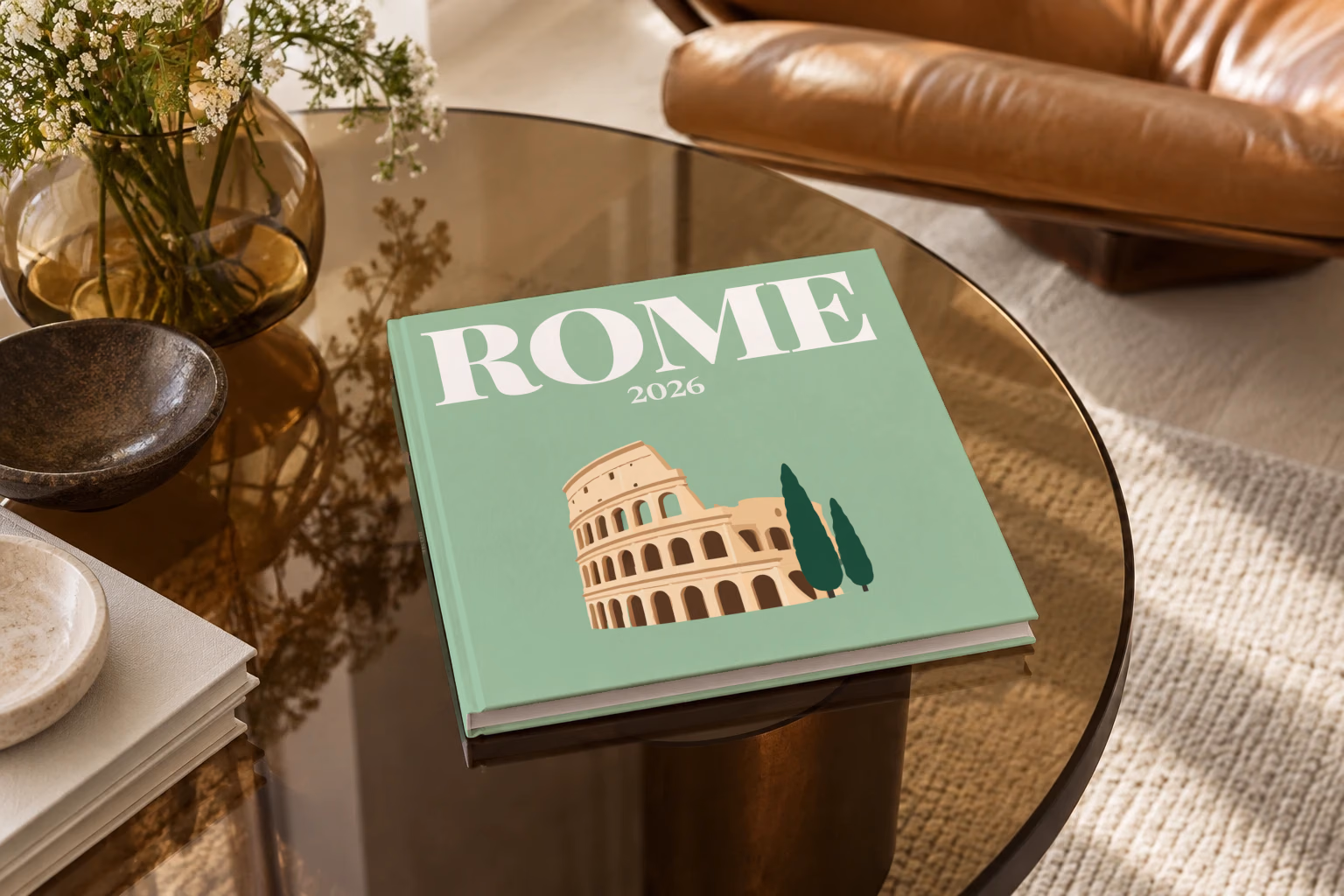 Italy square hardcover photo book with the Rome illustrated cover of the Colosseum and cypress trees on sage green, on a smoked glass coffee table beside a leather armchair in warm afternoon light