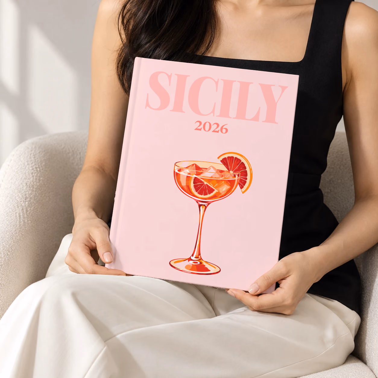 Italy hardcover photo book with the Sicily illustrated cover of a blood orange spritz on pale pink, held upright in two hands by a woman in a black top, sitting in a cream boucle armchair