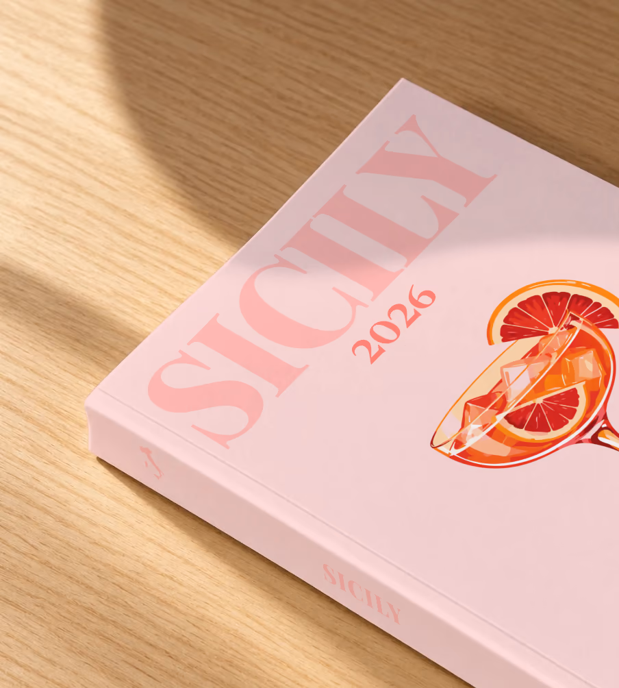Italy softcover photo book with the Sicily illustrated cover of a blood orange spritz on pale pink, lying on an oak desk in dappled afternoon sunlight