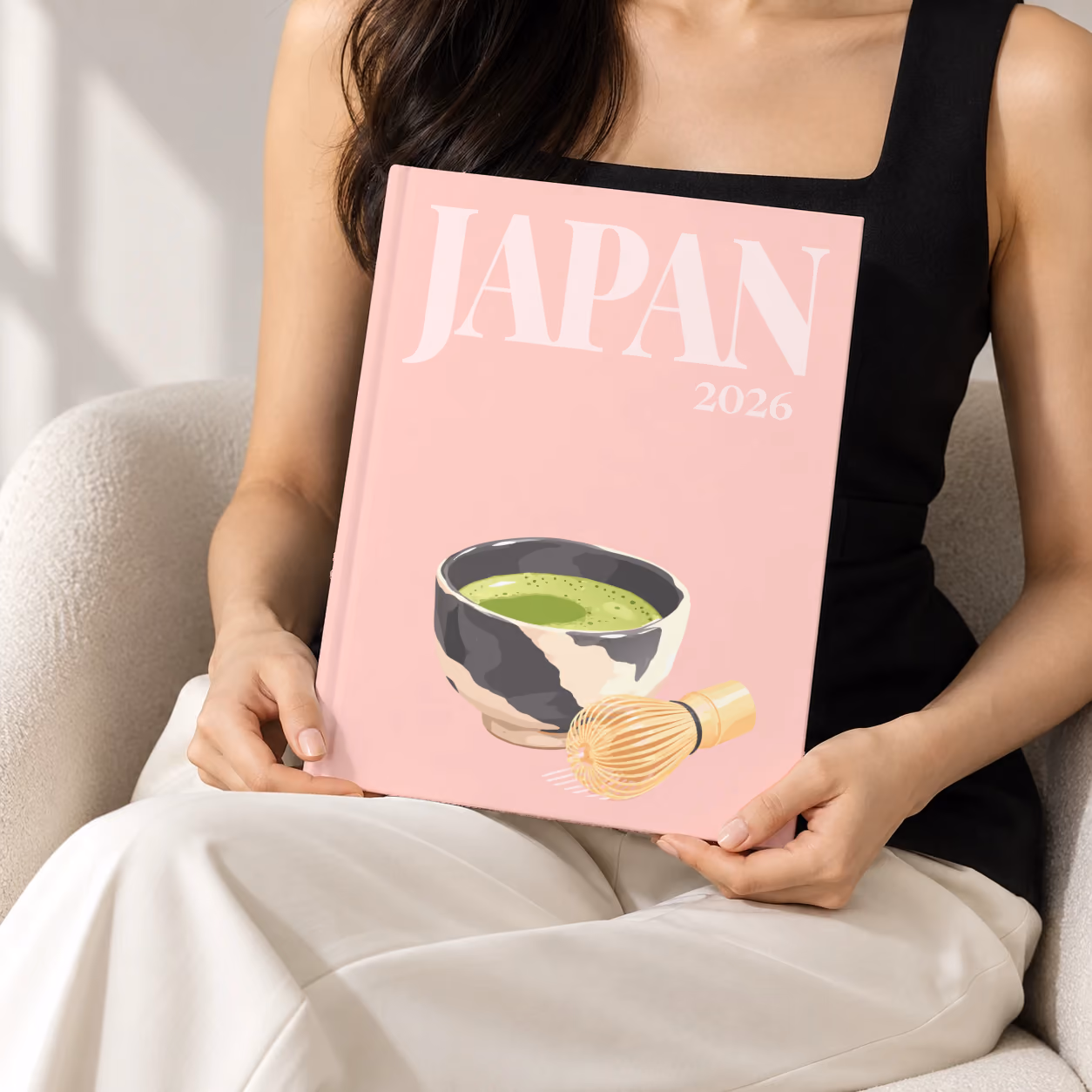 Japan hardcover photo book with the illustrated cover of a bowl of matcha and a whisk on pale pink, held upright in two hands by a woman in a black top, sitting in a cream boucle armchair