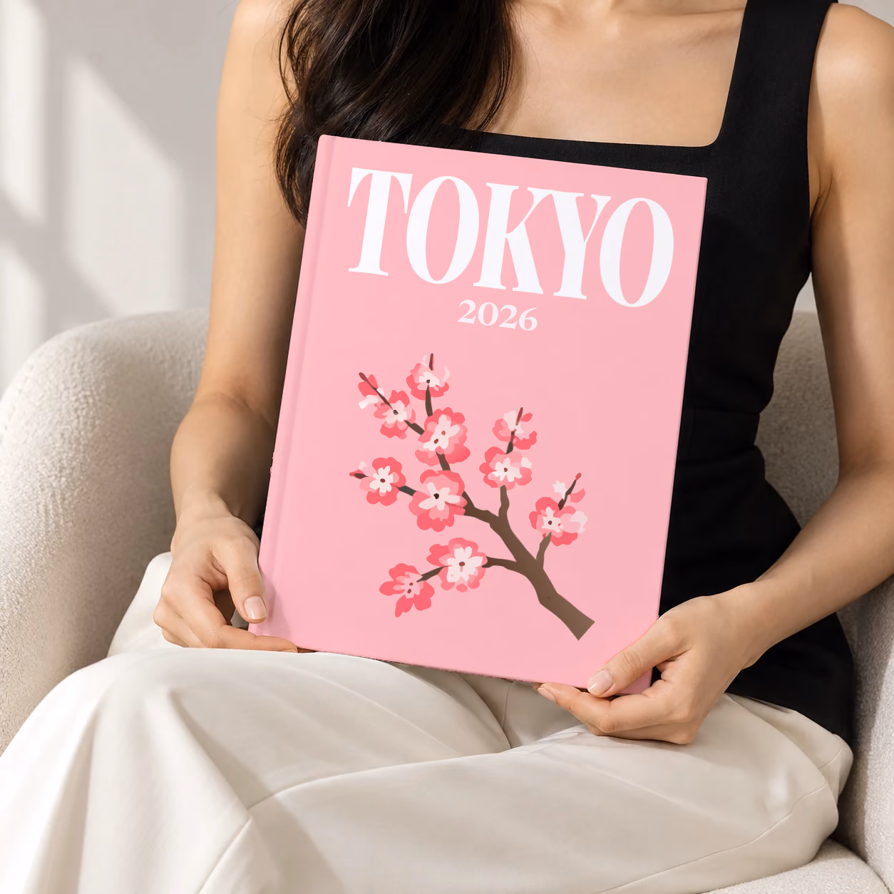 Japan hardcover photo book with the Tokyo illustrated cover of a cherry blossom branch on pink, held upright in two hands by a woman in a black top, sitting in a cream boucle armchair