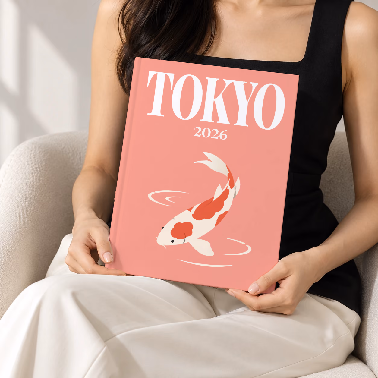 Japan hardcover photo book with the Tokyo illustrated cover of a koi on coral pink, held upright in two hands by a woman in a black top, sitting in a cream boucle armchair