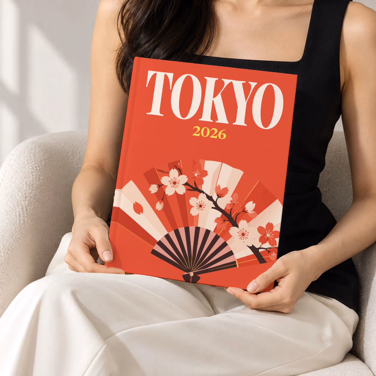 Japan hardcover photo book with the Tokyo illustrated cover of a folding fan with cherry blossom on red, held upright in two hands by a woman in a black top, sitting in a cream boucle armchair