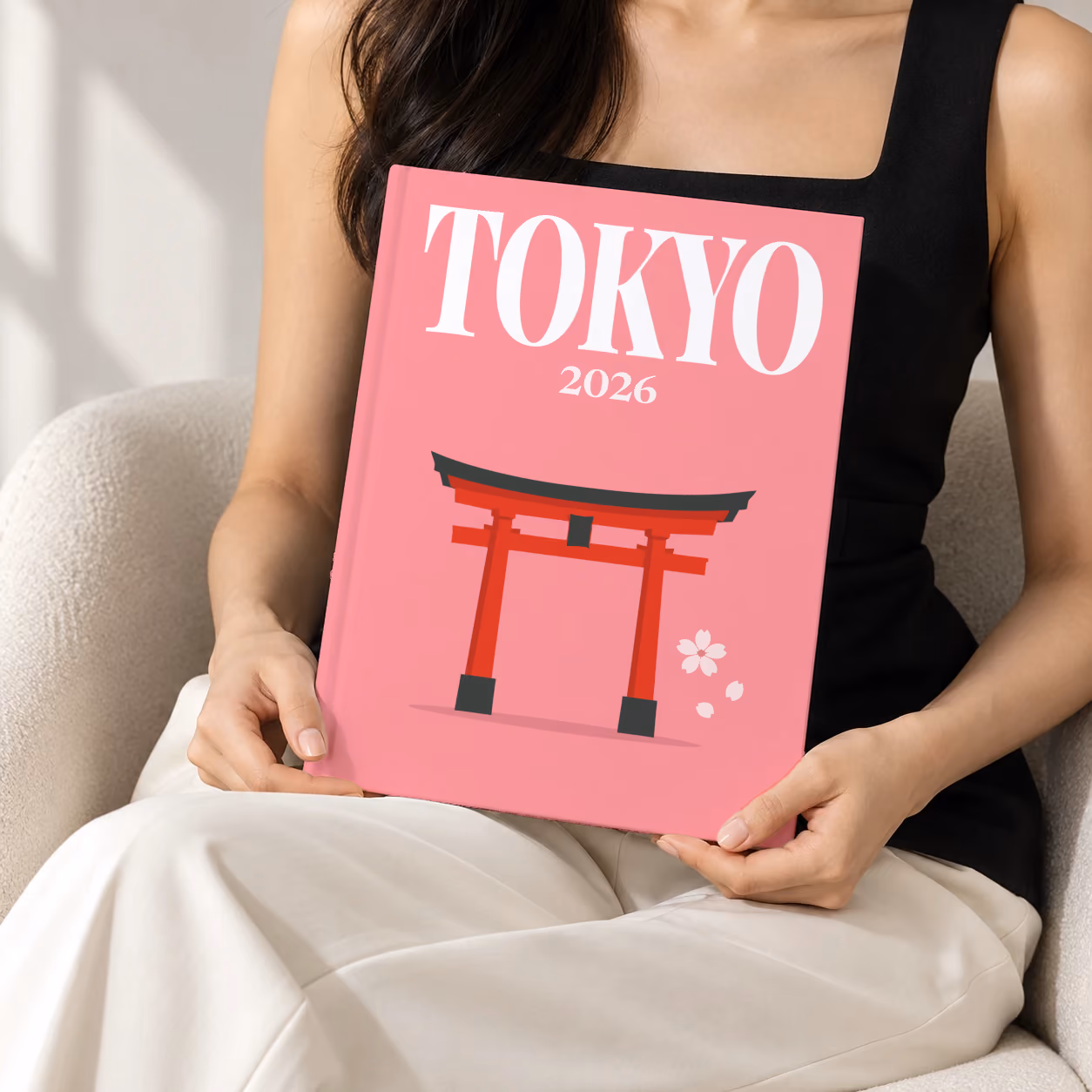 Japan hardcover photo book with the Tokyo illustrated cover of a red torii gate and petals on pink, held upright in two hands by a woman in a black top, sitting in a cream boucle armchair