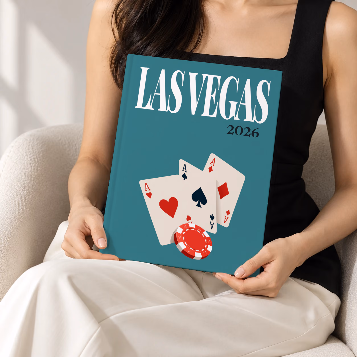 Las Vegas hardcover photo book with the illustrated cover of playing card aces and a red casino chip on teal, held upright in two hands by a woman in a black top, sitting in a cream boucle armchair