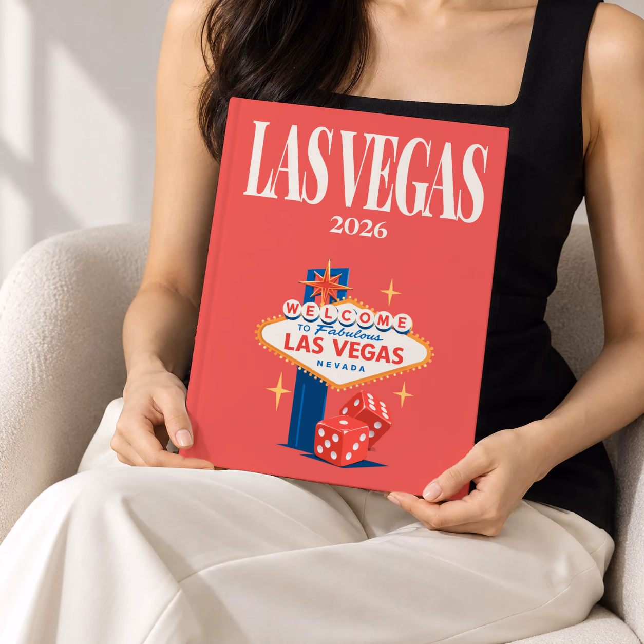 Las Vegas hardcover photo book with the illustrated cover of the Welcome to Fabulous Las Vegas sign and red dice on red, held upright in two hands by a woman in a black top, sitting in a cream boucle armchair