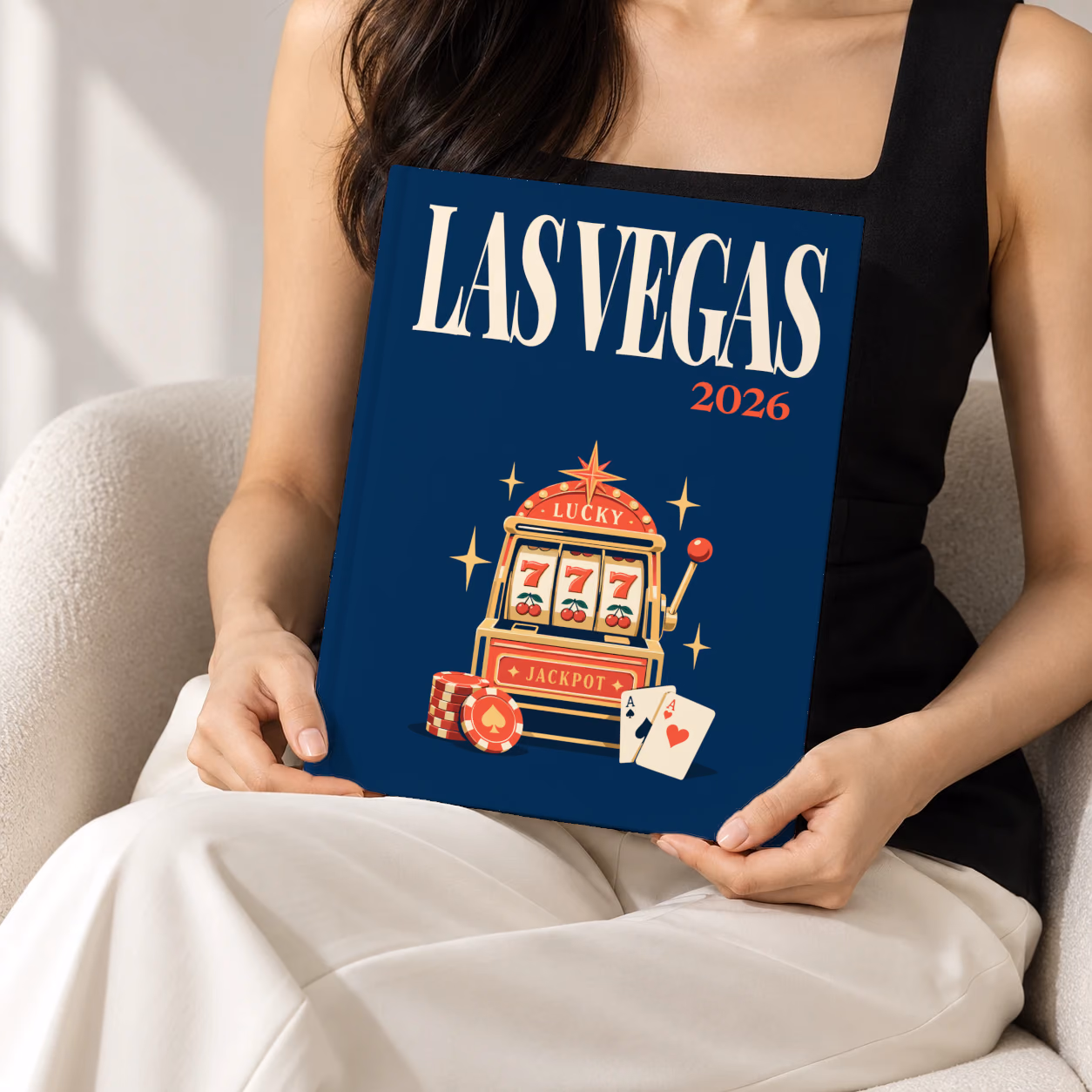 Las Vegas hardcover photo book with the illustrated cover of a lucky 777 jackpot slot machine with casino chips and cards on navy, held upright in two hands by a woman in a black top, sitting in a cream boucle armchair