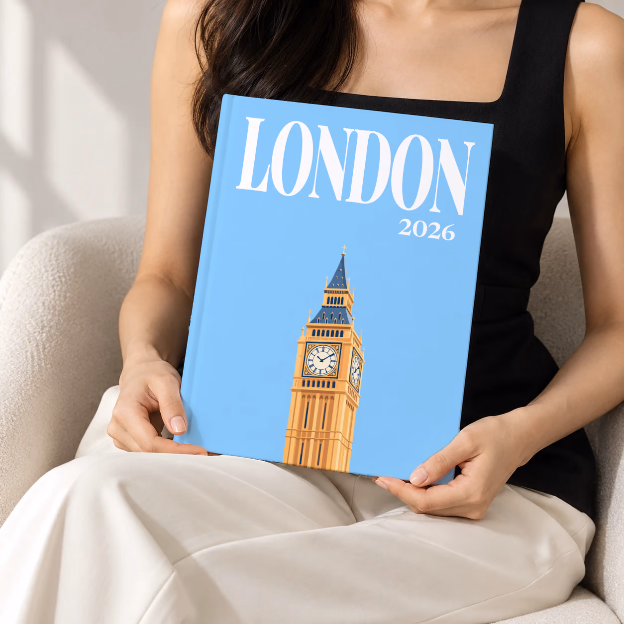 London hardcover photo book with the illustrated cover of Big Ben's Elizabeth Tower on sky blue, held upright in two hands by a woman in a black top, sitting in a cream boucle armchair