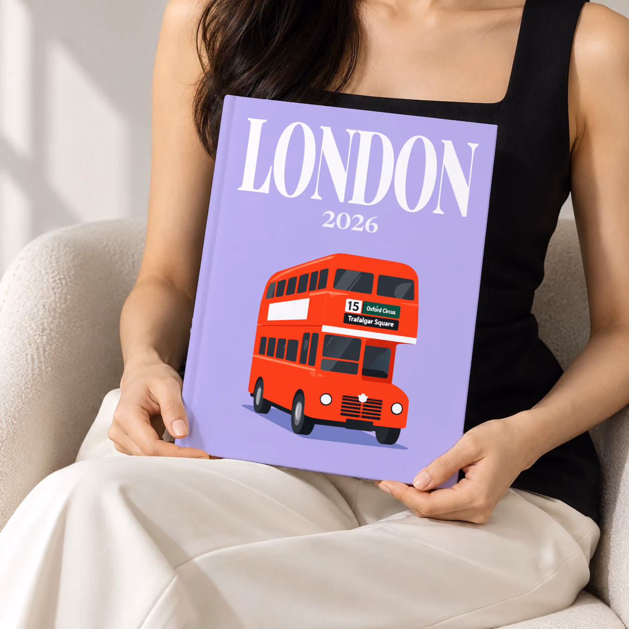 London hardcover photo book with the illustrated cover of a red double-decker London bus to Oxford Circus and Trafalgar Square on lavender, held upright in two hands by a woman in a black top, sitting in a cream boucle armchair
