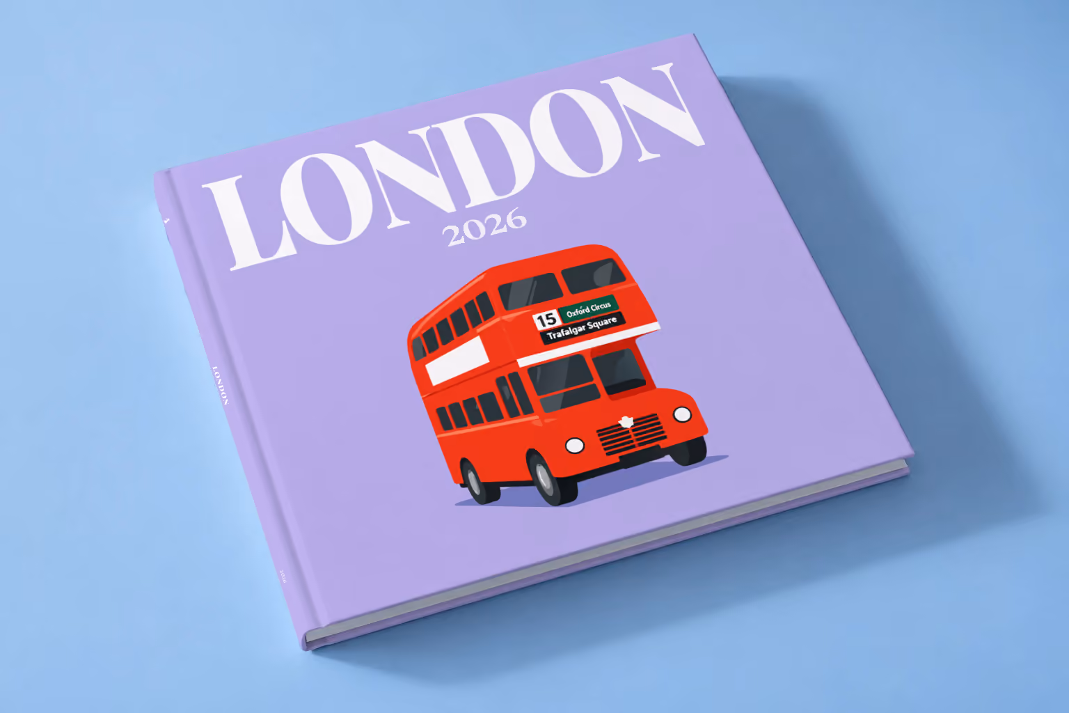 London square hardcover photo book with the illustrated cover of a red double-decker London bus to Oxford Circus and Trafalgar Square on lavender, lying on a pale blue studio backdrop
