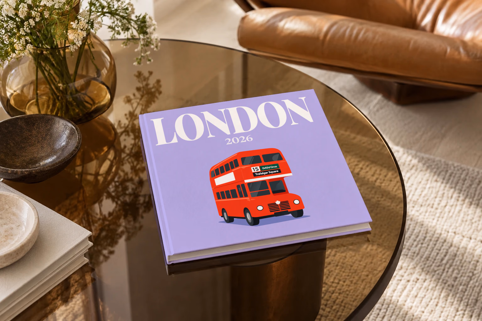 London square hardcover photo book with the illustrated cover of a red double-decker London bus to Oxford Circus and Trafalgar Square on lavender, on a smoked glass coffee table beside a leather armchair in warm afternoon light
