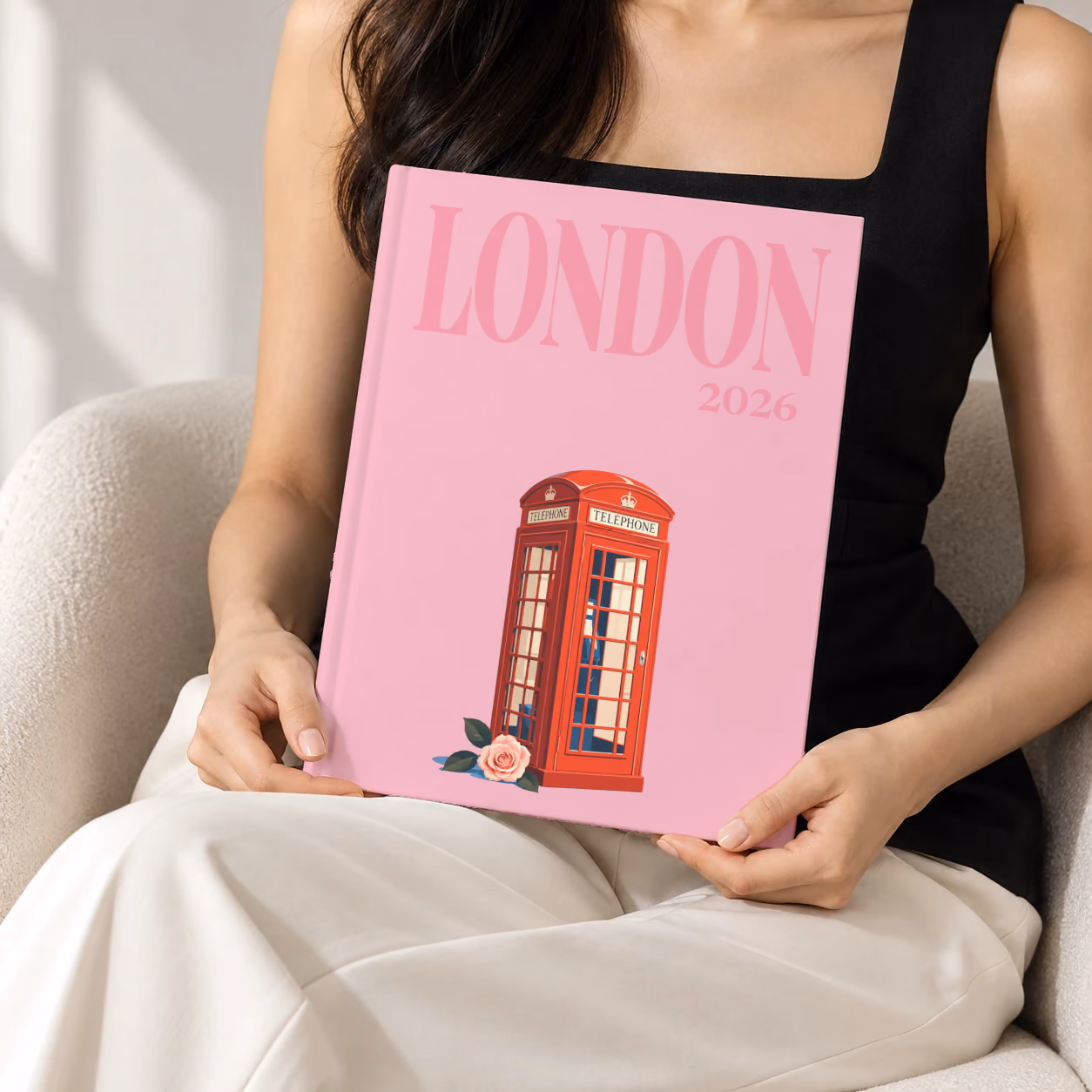 London hardcover photo book with the illustrated cover of a red London telephone box and a pink rose on pink, held upright in two hands by a woman in a black top, sitting in a cream boucle armchair