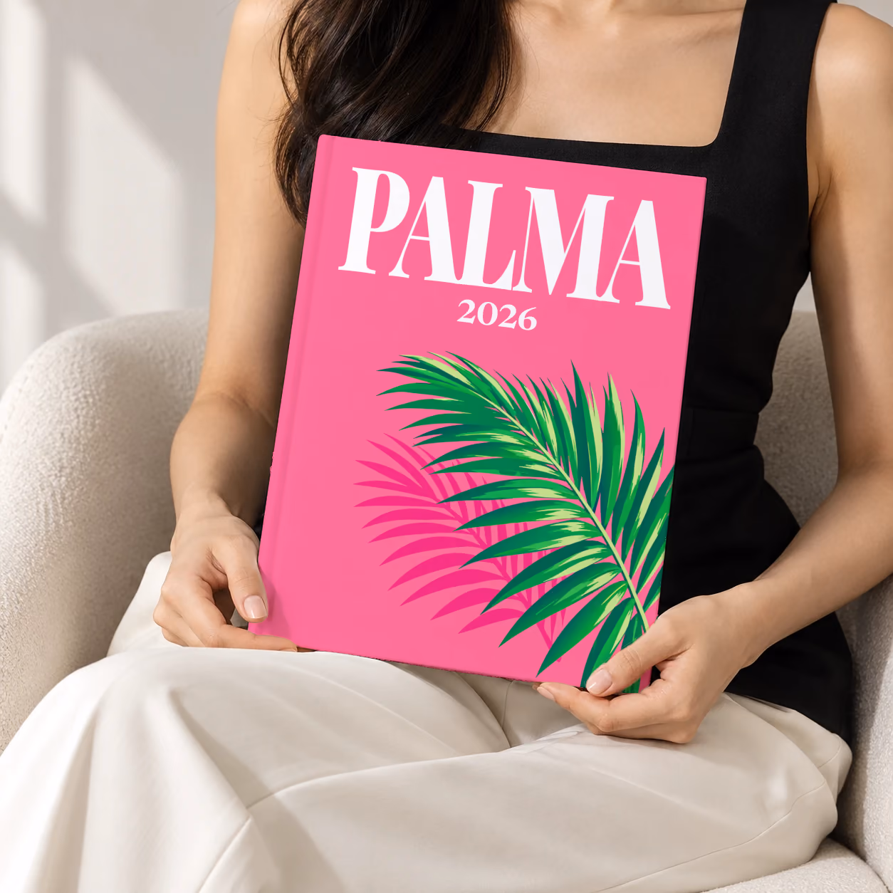 Mallorca hardcover photo book with the Palma illustrated cover of a palm frond on hot pink, held upright in two hands by a woman in a black top, sitting in a cream boucle armchair