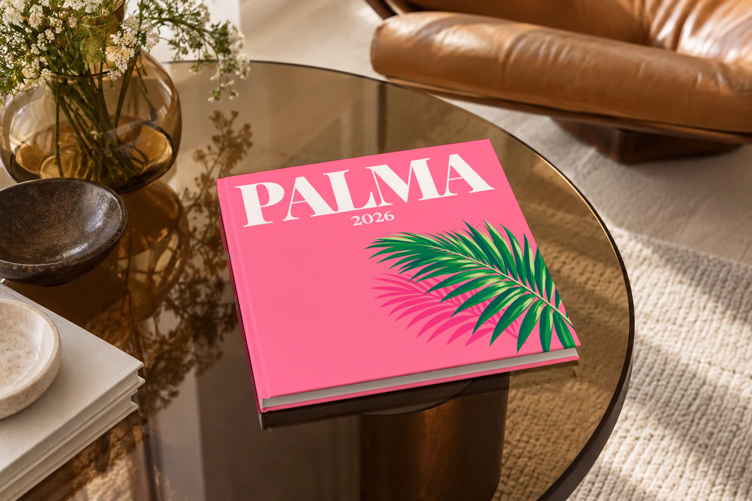 Mallorca square hardcover photo book with the Palma illustrated cover of a palm frond on hot pink, on a smoked glass coffee table beside a leather armchair in warm afternoon light