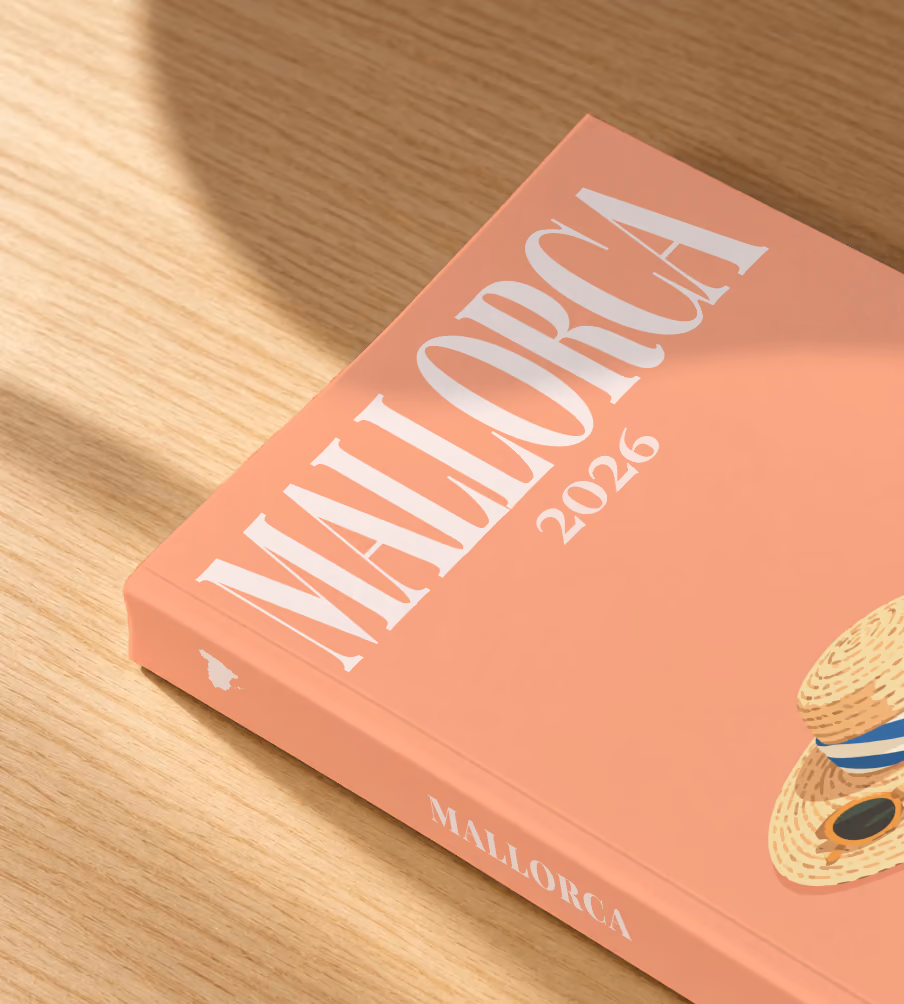 Mallorca softcover photo book with the illustrated cover of a straw hat with a striped scarf and a lemon on peach, lying on an oak desk in dappled afternoon sunlight