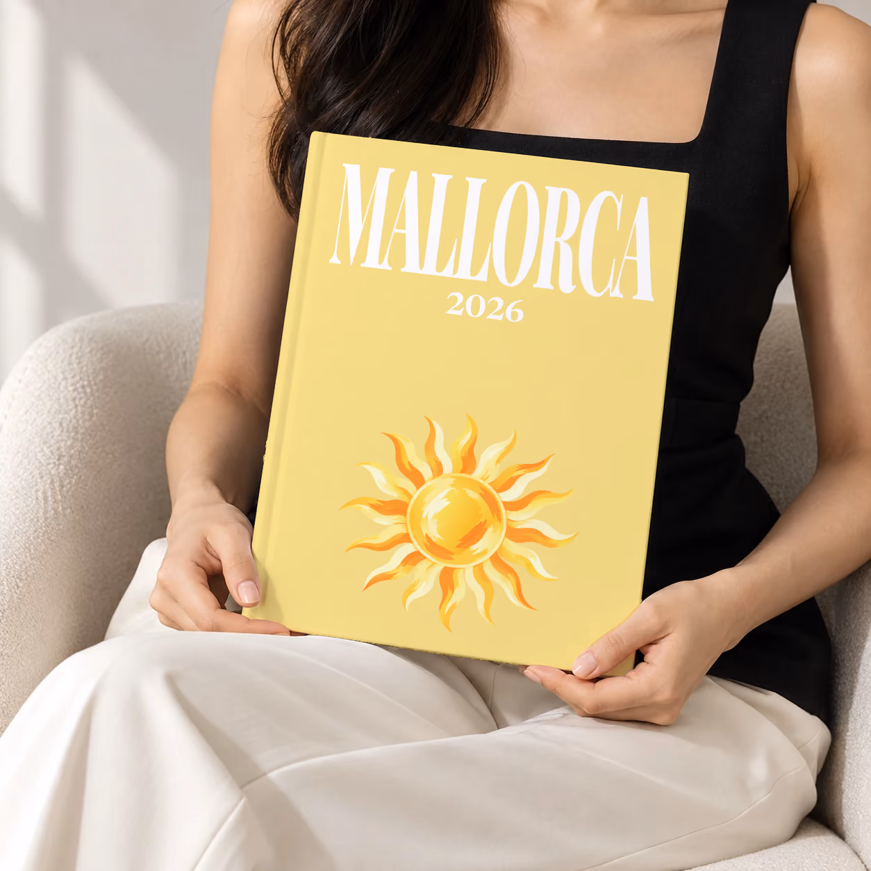 Mallorca hardcover photo book with the illustrated cover of a golden sun on pale yellow, held upright in two hands by a woman in a black top, sitting in a cream boucle armchair