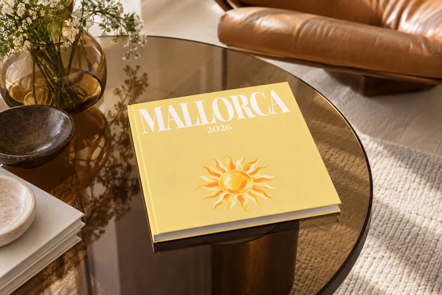 Mallorca square hardcover photo book with the illustrated cover of a golden sun on pale yellow, on a smoked glass coffee table beside a leather armchair in warm afternoon light