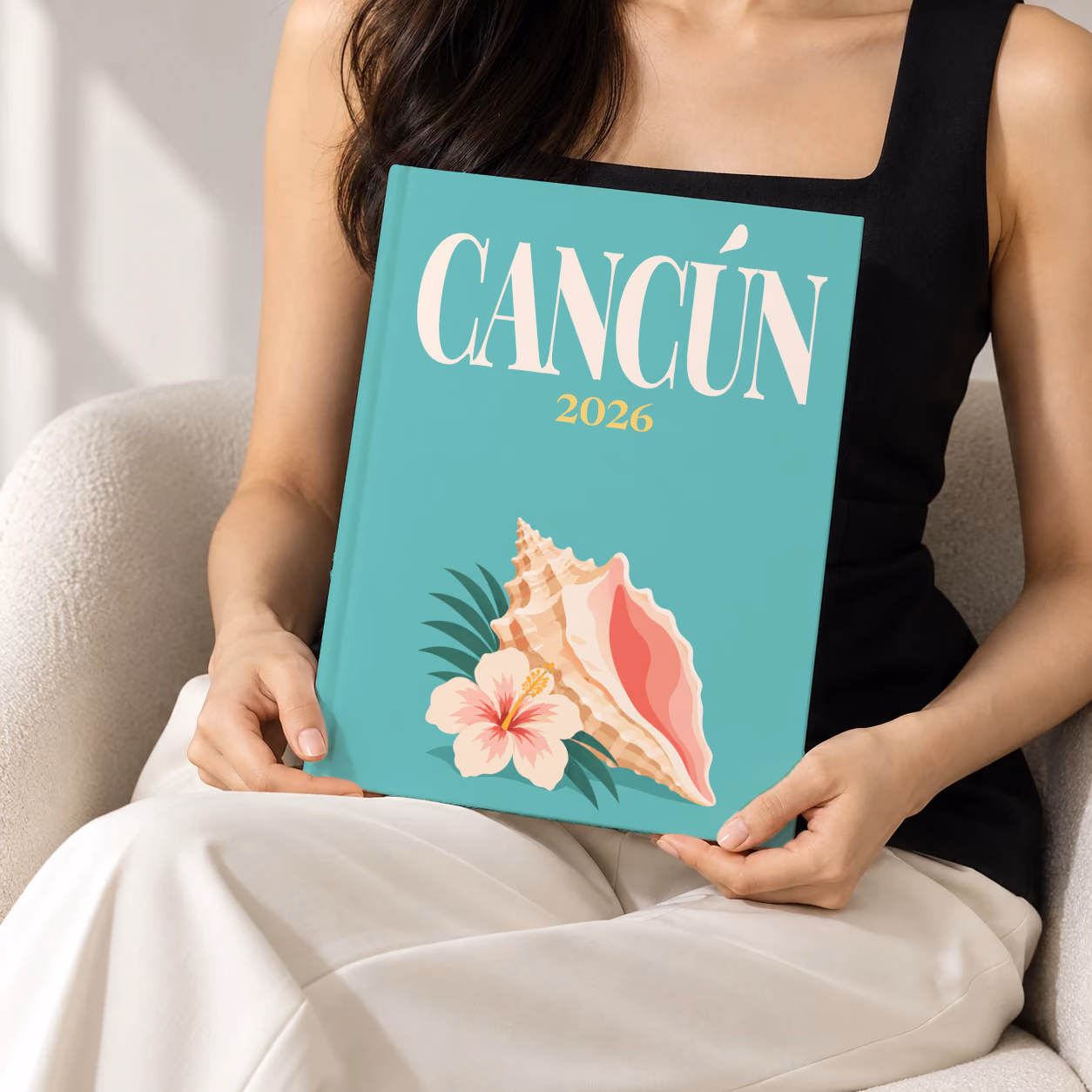 Mexico hardcover photo book with the Cancún illustrated cover of a conch shell and hibiscus on teal, held upright in two hands by a woman in a black top, sitting in a cream boucle armchair