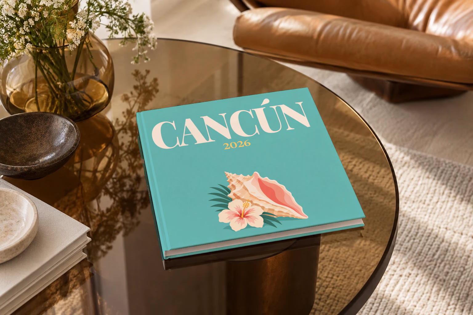 Mexico square hardcover photo book with the Cancún illustrated cover of a conch shell and hibiscus on teal, on a smoked glass coffee table beside a leather armchair in warm afternoon light