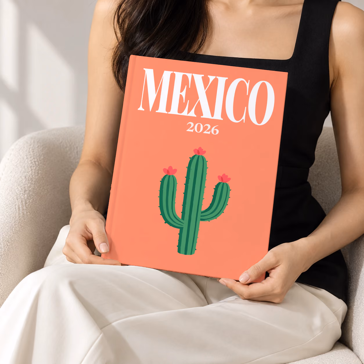 Mexico hardcover photo book with the illustrated cover of a flowering cactus on coral, held upright in two hands by a woman in a black top, sitting in a cream boucle armchair