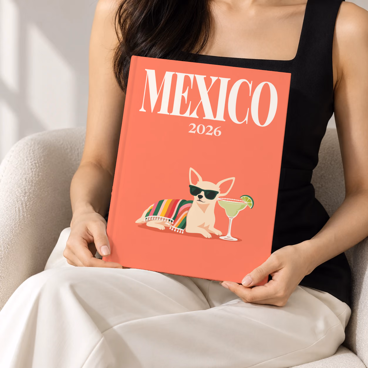 Mexico hardcover photo book with the illustrated cover of a chihuahua in sunglasses with a margarita on coral, held upright in two hands by a woman in a black top, sitting in a cream boucle armchair