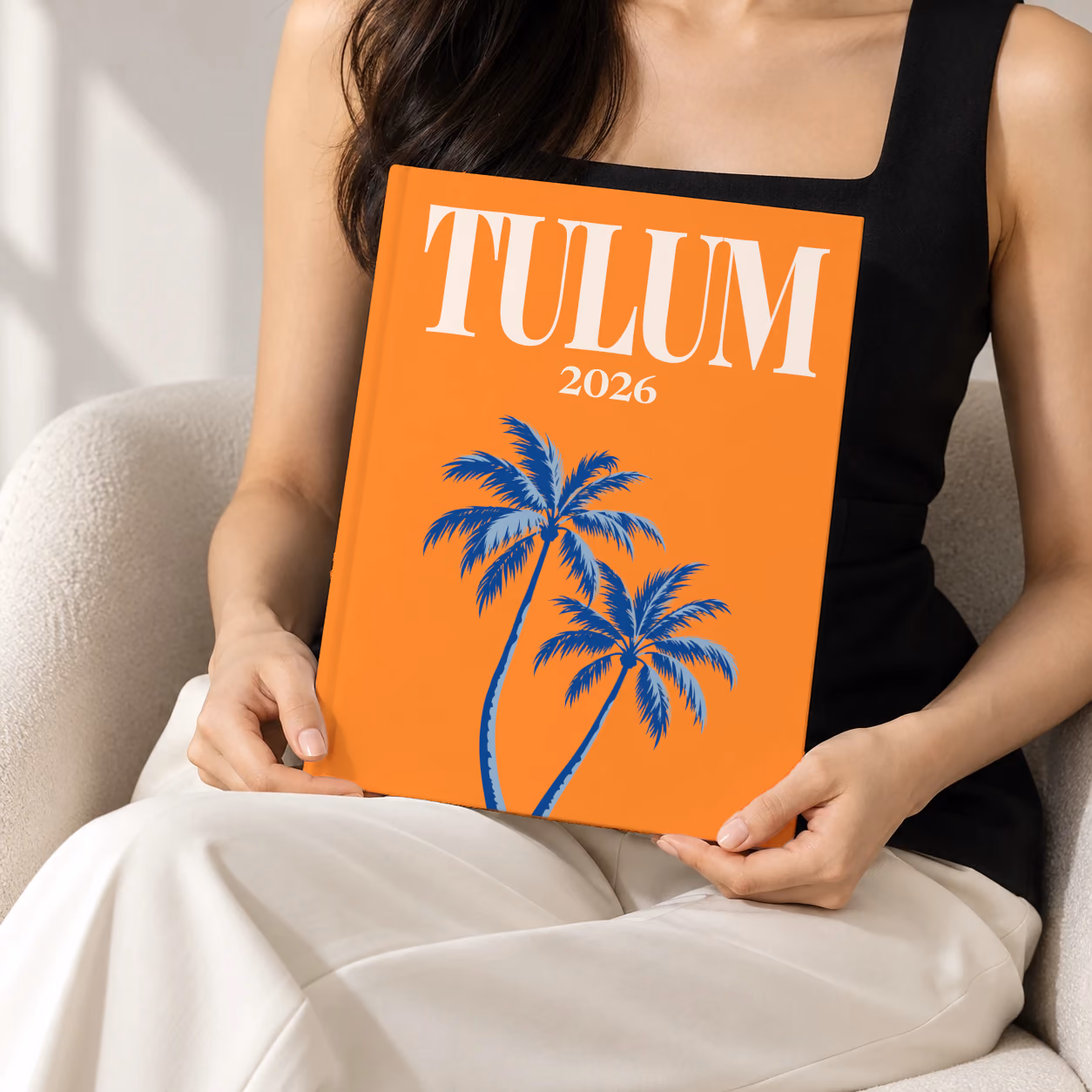 Mexico hardcover photo book with the Tulum illustrated cover of palm trees on orange, held upright in two hands by a woman in a black top, sitting in a cream boucle armchair