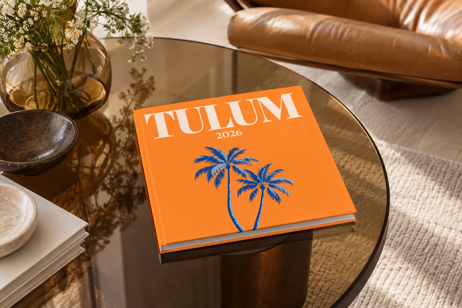 Mexico square hardcover photo book with the Tulum illustrated cover of palm trees on orange, on a smoked glass coffee table beside a leather armchair in warm afternoon light
