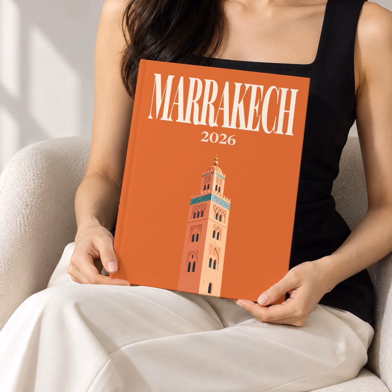 Morocco hardcover photo book with the Marrakech illustrated cover of the Koutoubia Mosque minaret on burnt orange, held upright in two hands by a woman in a black top, sitting in a cream boucle armchair