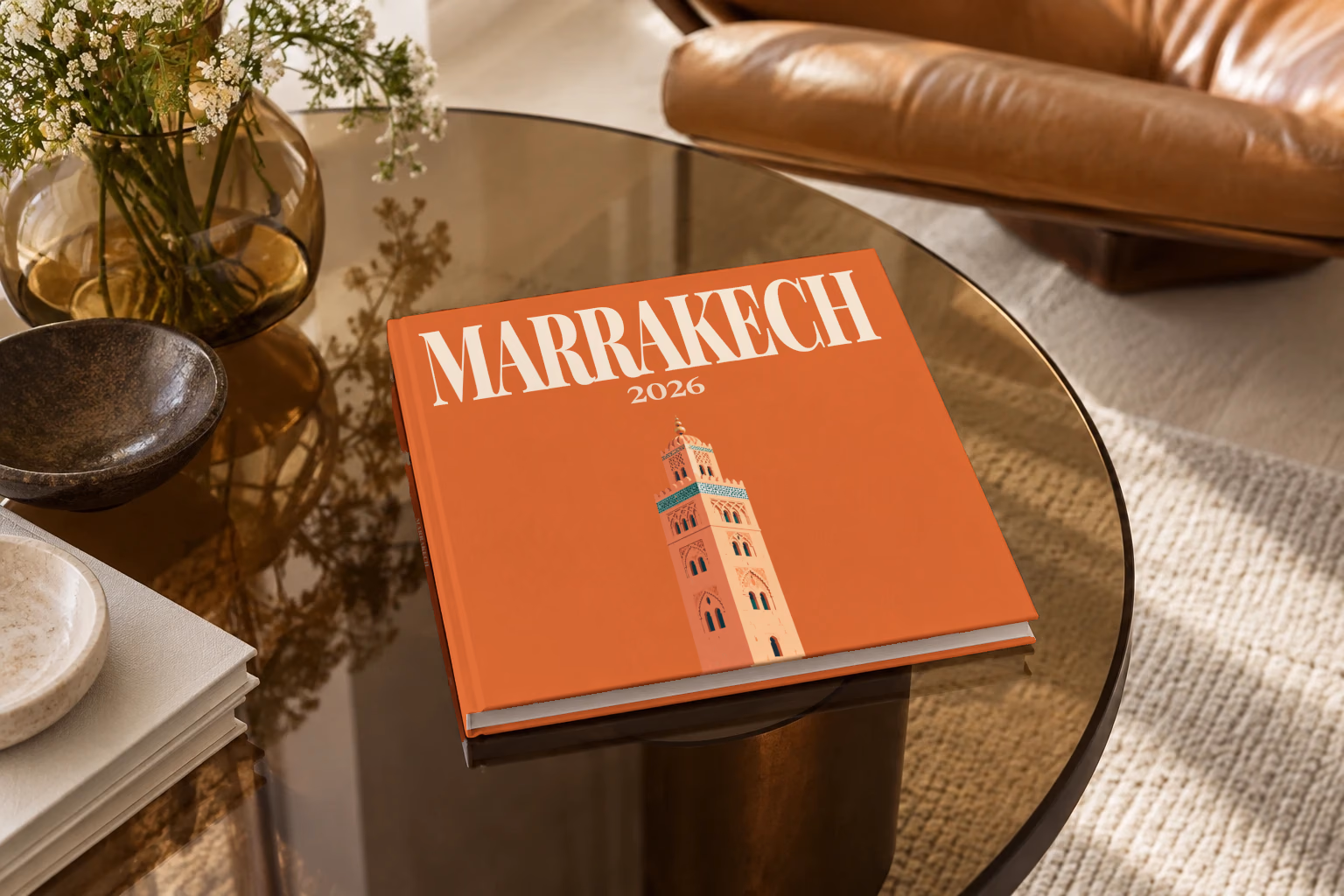 Morocco square hardcover photo book with the Marrakech illustrated cover of the Koutoubia Mosque minaret on burnt orange, on a smoked glass coffee table beside a leather armchair in warm afternoon light