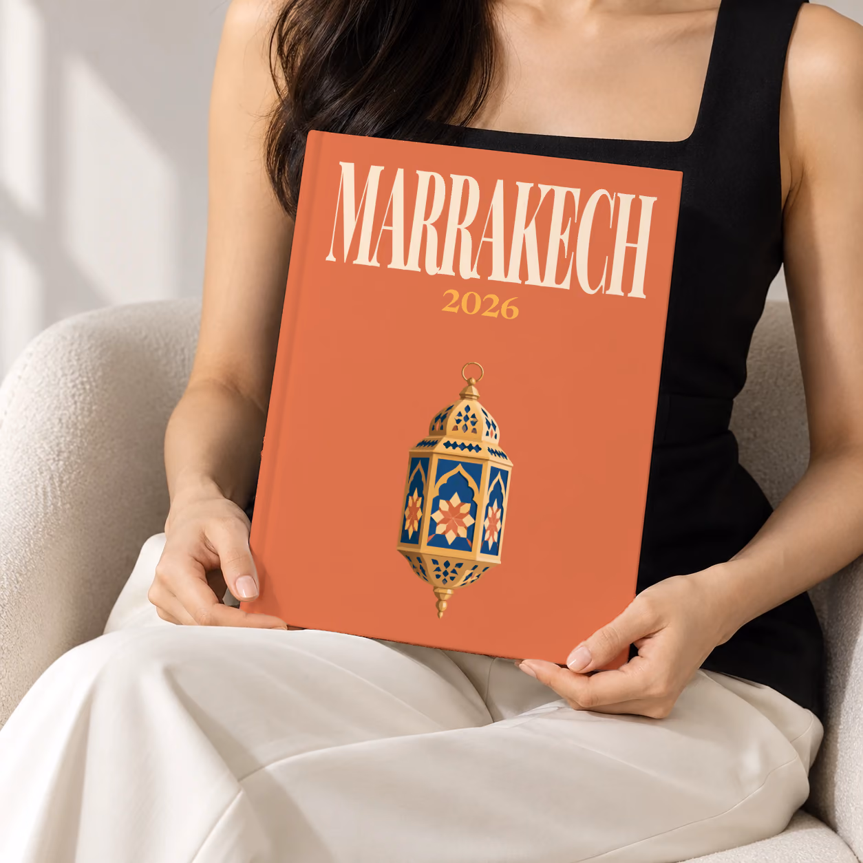 Morocco hardcover photo book with the Marrakech illustrated cover of a gold and blue Moroccan lantern on terracotta, held upright in two hands by a woman in a black top, sitting in a cream boucle armchair