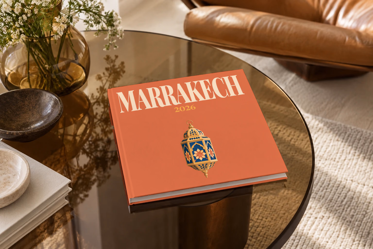 Morocco square hardcover photo book with the Marrakech illustrated cover of a gold and blue Moroccan lantern on terracotta, on a smoked glass coffee table beside a leather armchair in warm afternoon light