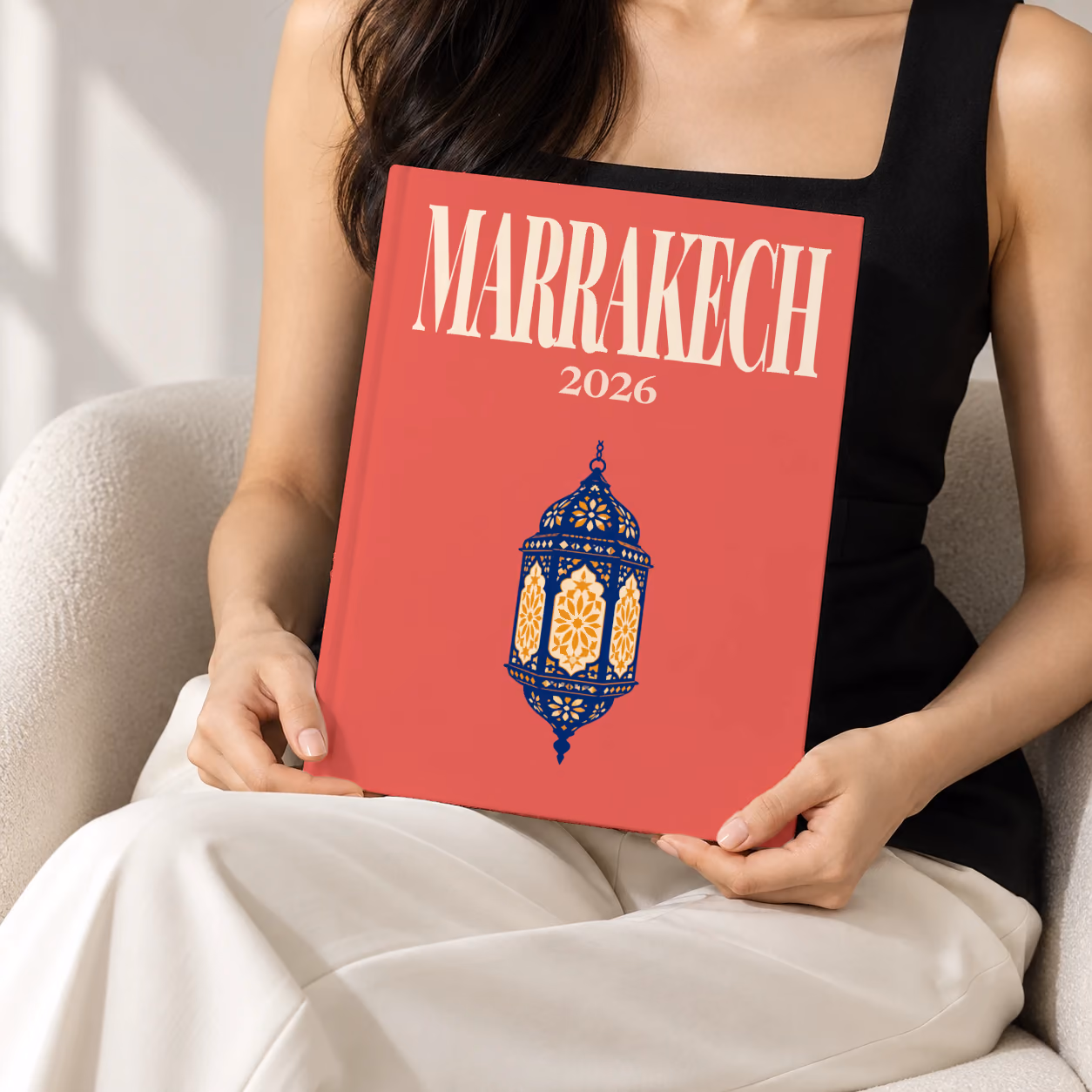 Morocco hardcover photo book with the Marrakech illustrated cover of a navy and gold Moroccan lantern on red, held upright in two hands by a woman in a black top, sitting in a cream boucle armchair