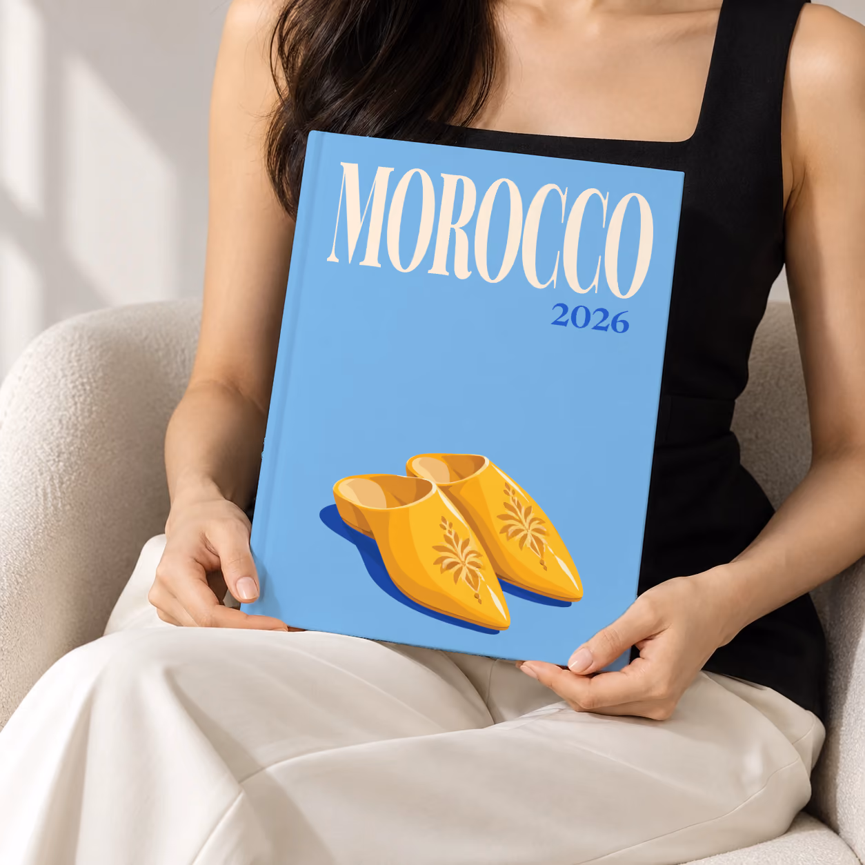 Morocco hardcover photo book with the illustrated cover of a pair of yellow Moroccan babouche slippers on sky blue, held upright in two hands by a woman in a black top, sitting in a cream boucle armchair