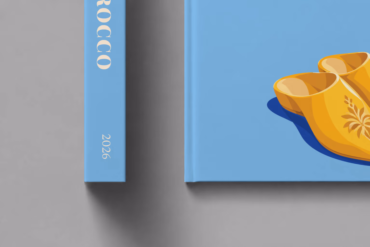 Morocco hardcover photo book with the illustrated cover of a pair of yellow Moroccan babouche slippers on sky blue, shown close up beside its spine on a pale grey studio surface