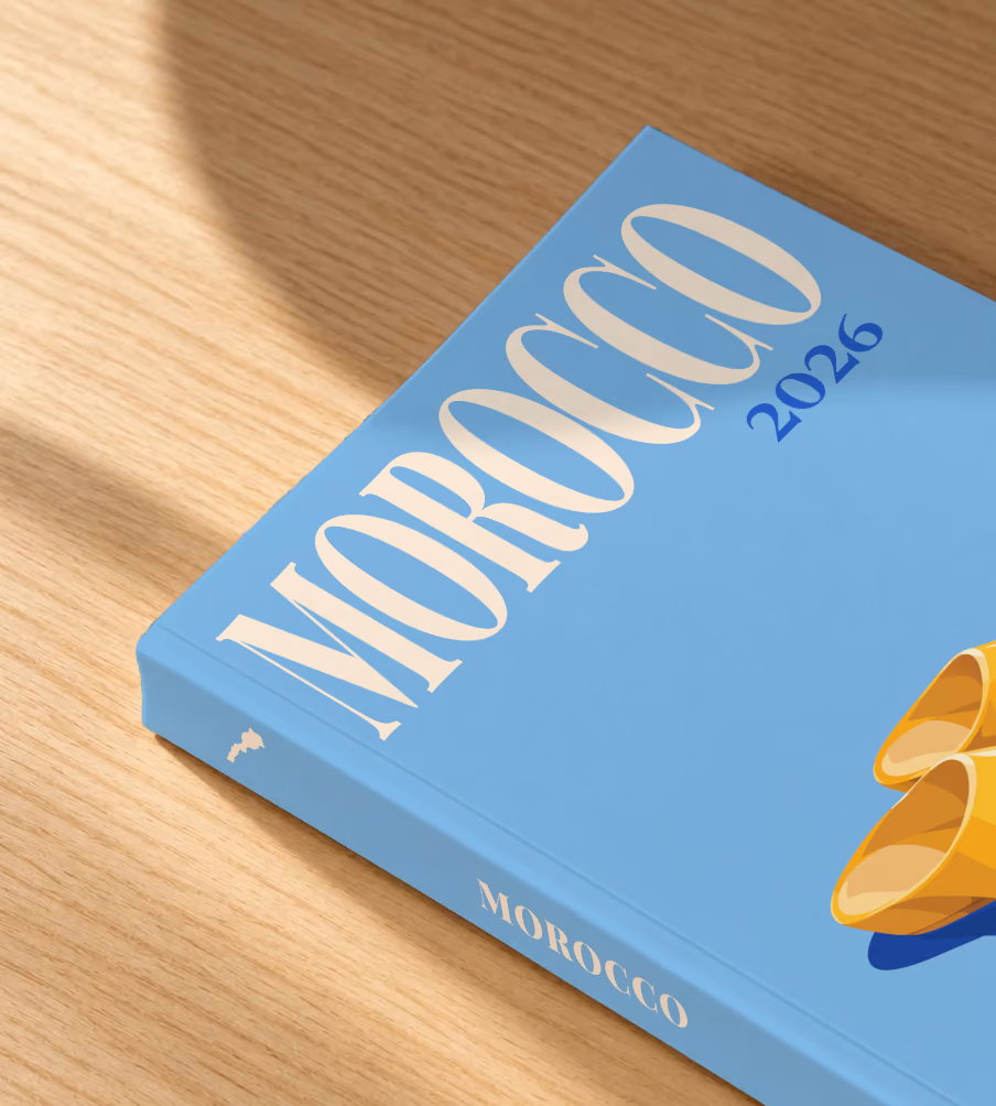 Morocco softcover photo book with the illustrated cover of a pair of yellow Moroccan babouche slippers on sky blue, lying on an oak desk in dappled afternoon sunlight