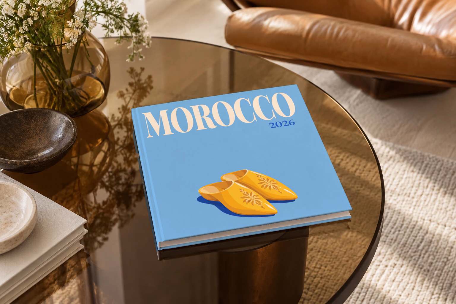 Morocco square hardcover photo book with the illustrated cover of a pair of yellow Moroccan babouche slippers on sky blue, on a smoked glass coffee table beside a leather armchair in warm afternoon light