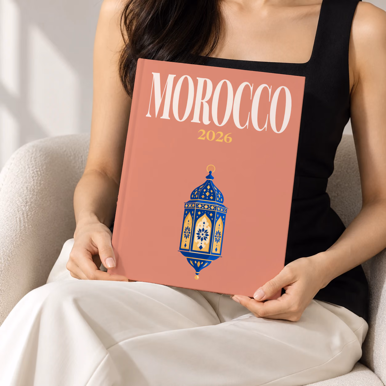 Morocco hardcover photo book with the illustrated cover of a blue and gold Moroccan lantern on dusty terracotta, held upright in two hands by a woman in a black top, sitting in a cream boucle armchair