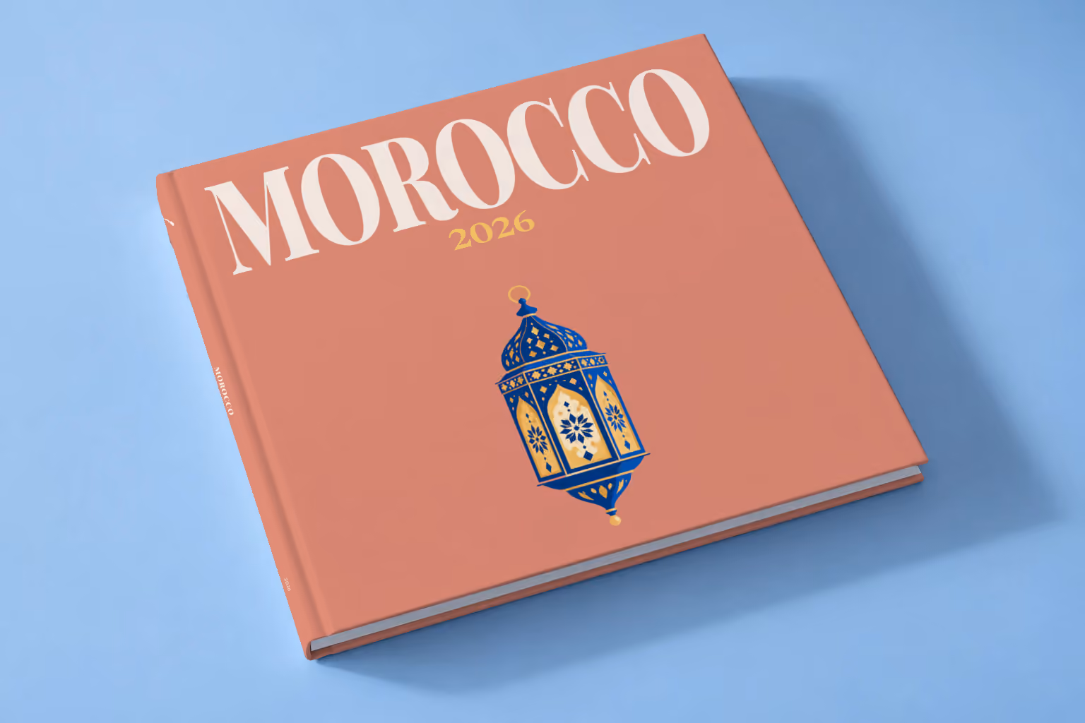 Morocco square hardcover photo book with the illustrated cover of a blue and gold Moroccan lantern on dusty terracotta, lying on a pale blue studio backdrop