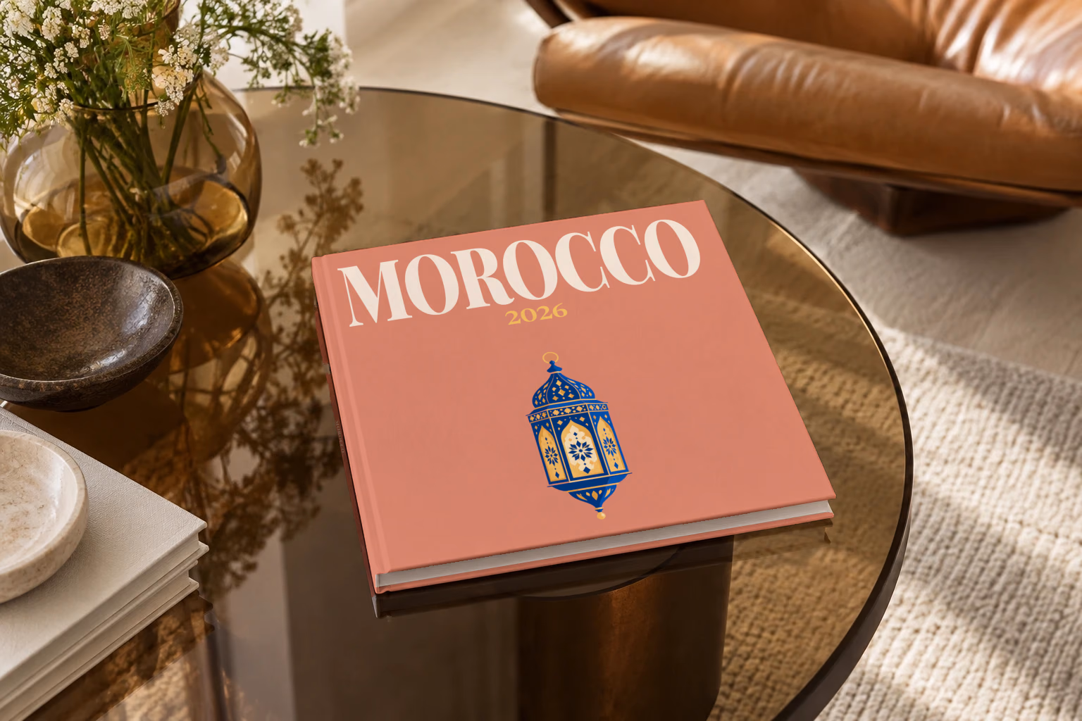 Morocco square hardcover photo book with the illustrated cover of a blue and gold Moroccan lantern on dusty terracotta, on a smoked glass coffee table beside a leather armchair in warm afternoon light