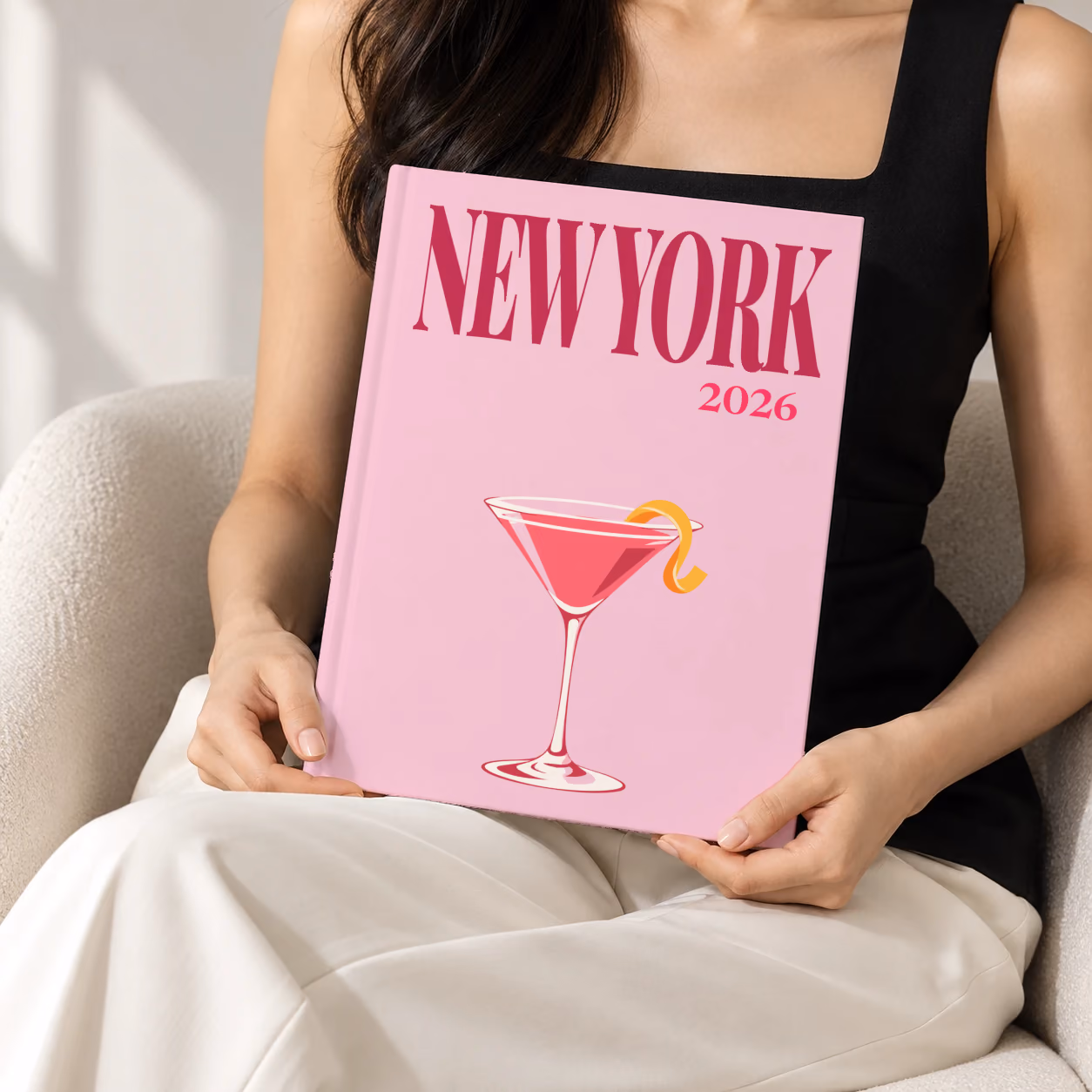 New York hardcover photo book with the illustrated cover of a cosmopolitan on pale pink, held upright in two hands by a woman in a black top, sitting in a cream boucle armchair
