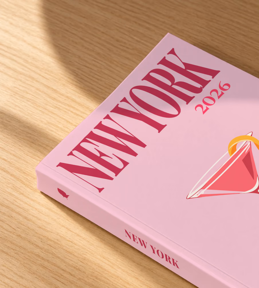 New York softcover photo book with the illustrated cover of a cosmopolitan on pale pink, lying on an oak desk in dappled afternoon sunlight