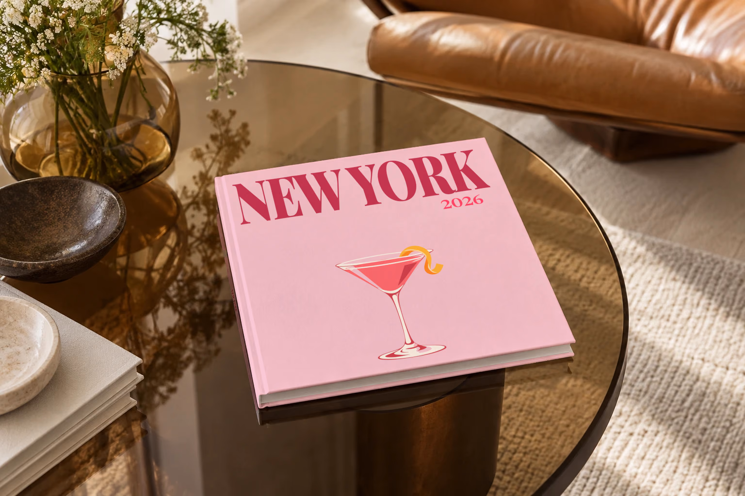 New York square hardcover photo book with the illustrated cover of a cosmopolitan on pale pink, on a smoked glass coffee table beside a leather armchair in warm afternoon light