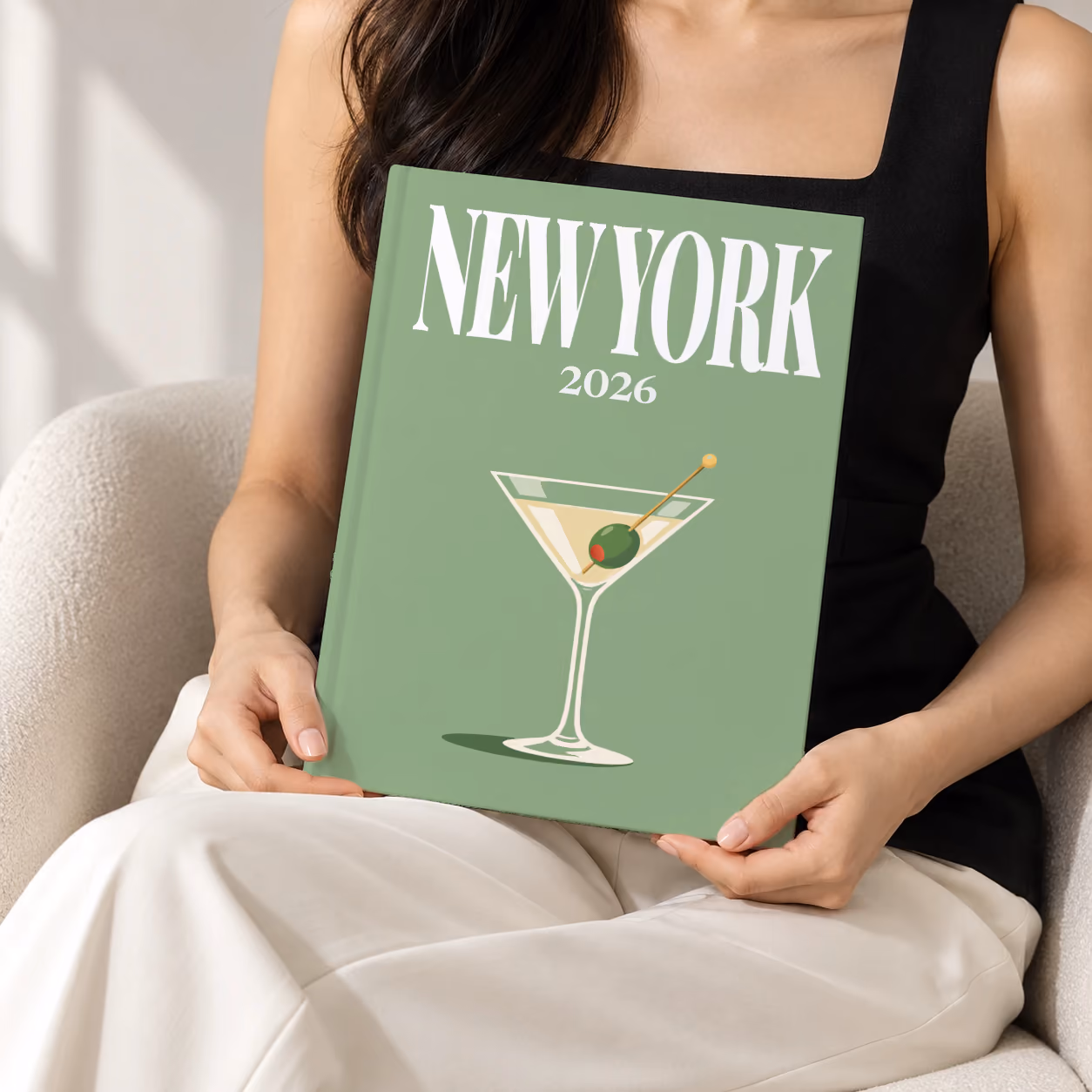 New York hardcover photo book with the illustrated cover of a martini with an olive on sage green, held upright in two hands by a woman in a black top, sitting in a cream boucle armchair