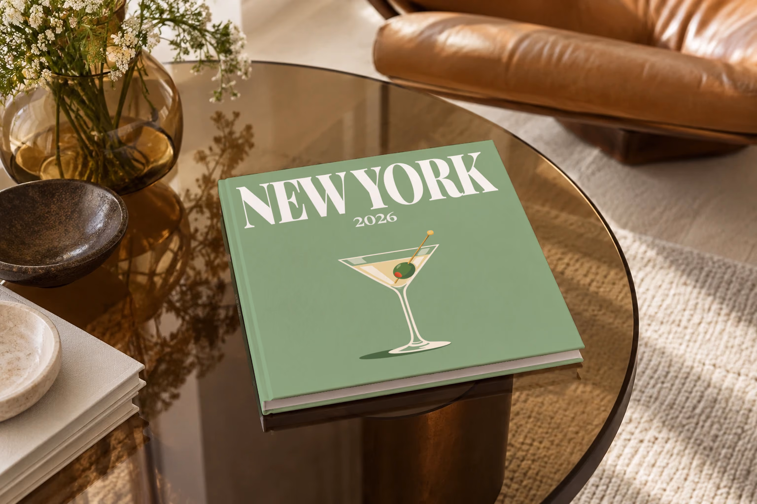 New York square hardcover photo book with the illustrated cover of a martini with an olive on sage green, on a smoked glass coffee table beside a leather armchair in warm afternoon light