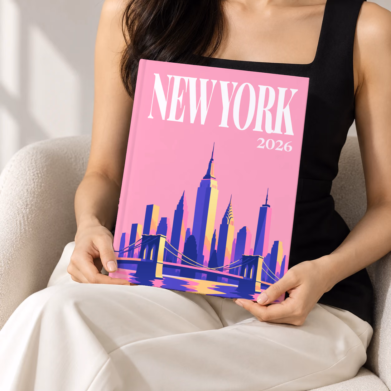 New York hardcover photo book with the illustrated cover of the Manhattan skyline and Brooklyn Bridge on pink, held upright in two hands by a woman in a black top, sitting in a cream boucle armchair
