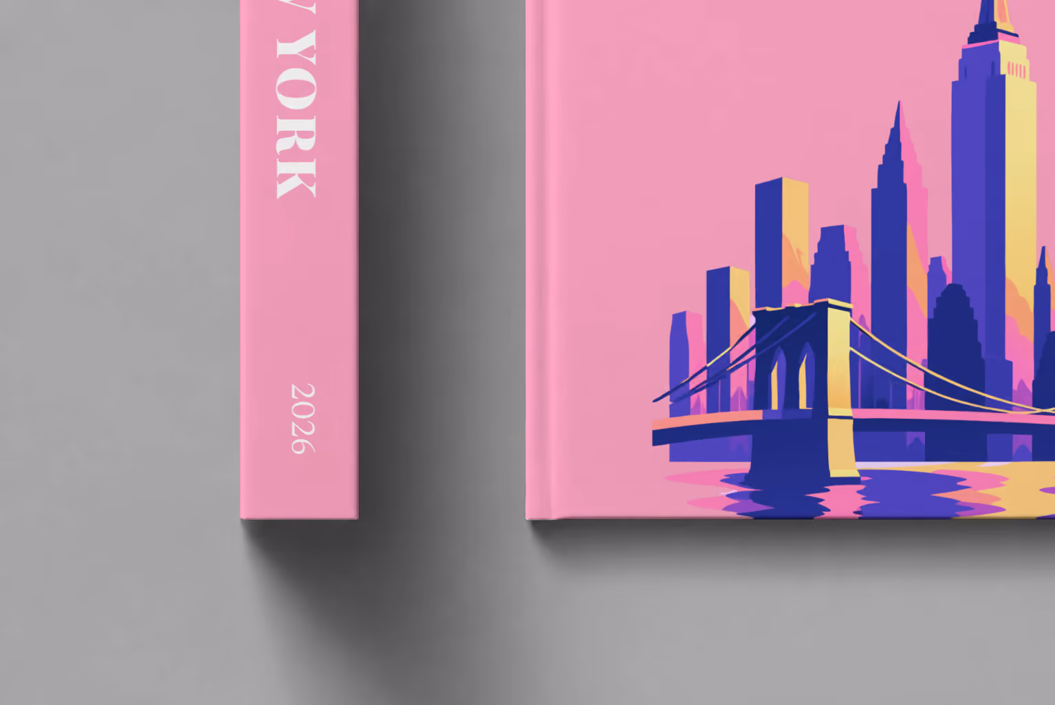 New York hardcover photo book with the illustrated cover of the Manhattan skyline and Brooklyn Bridge on pink, shown close up beside its spine on a pale grey studio surface