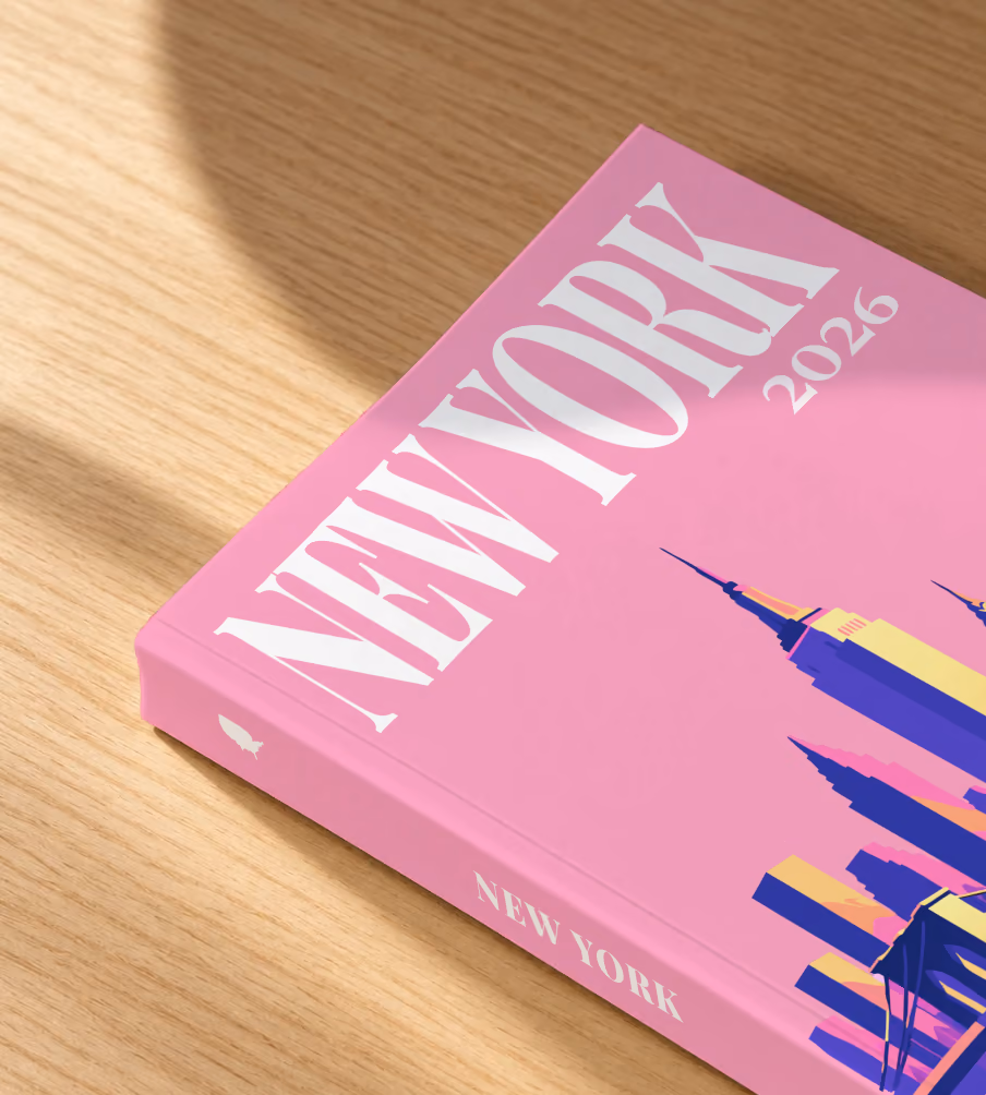 New York softcover photo book with the illustrated cover of the Manhattan skyline and Brooklyn Bridge on pink, lying on an oak desk in dappled afternoon sunlight